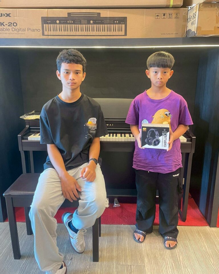 Two boys pose by a piano in a store; the left boy wears a black T‑shirt and light pants, the right boy in a purple shirt holds a headphone box.