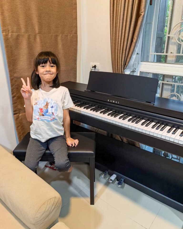 A young girl sits on a black piano bench beside a Kawai digital piano, flashing a peace sign at the camera.
