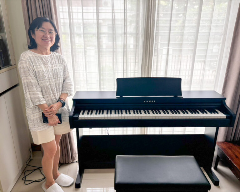 Smiling woman standing beside a black Kawai digital piano in a bright living room with sheer curtains behind her.