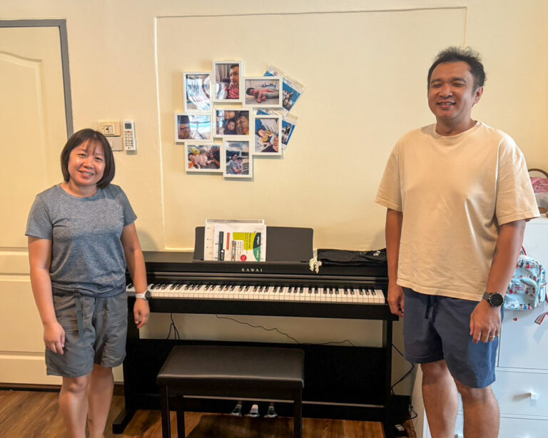 Two people stand beside a black Kawai upright piano in a cozy room, with a collage of framed photos on the wall behind them.