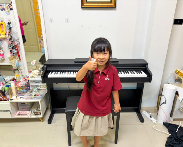 Young girl in a red polo gives a thumbs up while standing at a black digital piano in a cluttered room filled with craft supplies on the left.