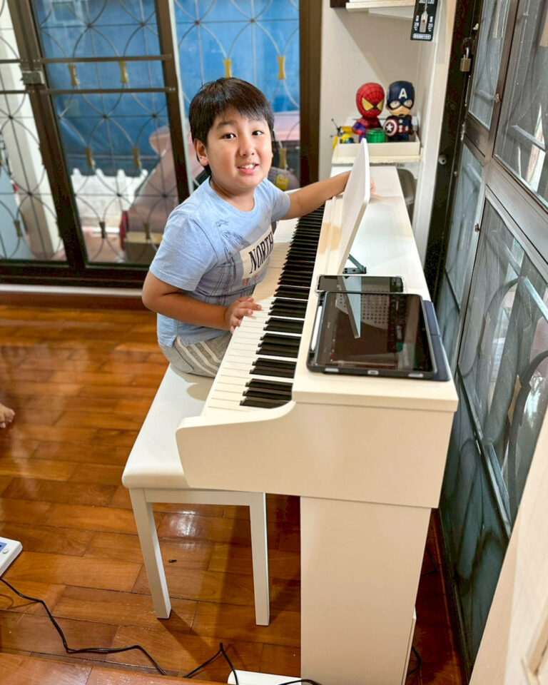 Young boy sits at a white piano, playing the keys with a tablet propped on the music stand nearby in a bright room with shelves.