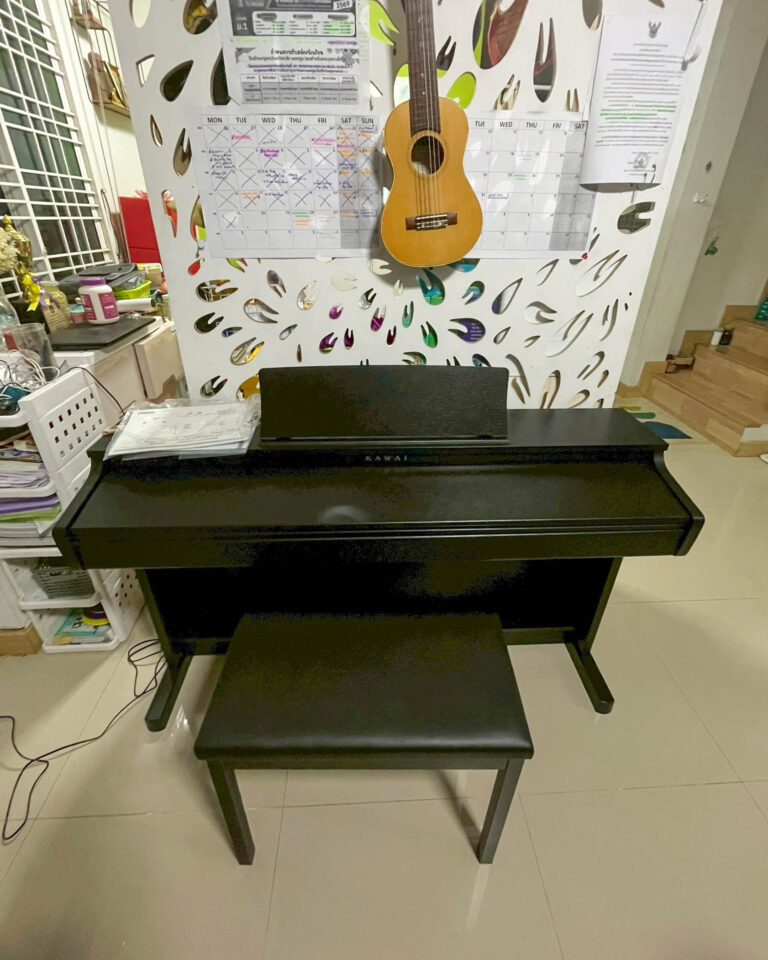 Black Kawai upright piano with matching bench in a cluttered room, calendar and papers on the wall behind, plus a small guitar hanging on a decorative panel.