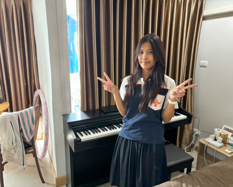 Young girl in a school uniform posing with peace signs beside a black piano in a cozy room with brown curtains.