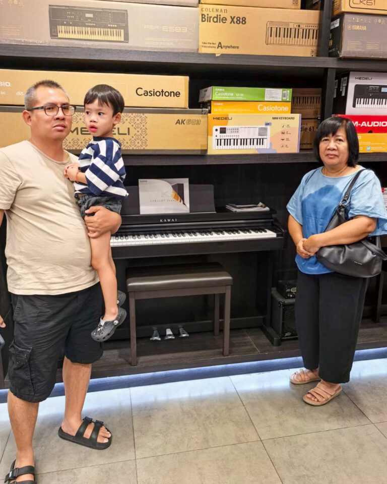 Man in a beige shirt holds a child beside a black digital piano in a music store, with boxed keyboards on shelves behind them.