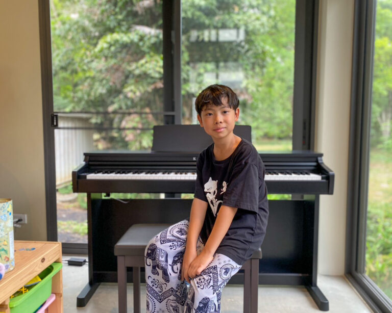 Boy sits on a piano bench in a sunlit room, positioned in front of a black upright piano beside large windows.