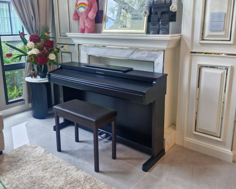 Black upright piano with bench in a bright living room, marble fireplace mantel above, decorative items and framed photo on the mantel, and a vase of red and white flowers on a side table by a large window.