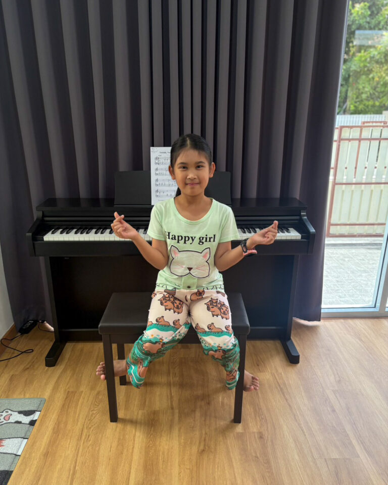 Young girl sits on a piano bench in front of a black upright piano, smiling at the camera with sheet music on the stand behind her.