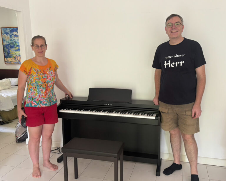 Two people stand beside a black Kawai digital piano in a bright room; woman on the left wears an orange floral top and red shorts, man on the right wears a black T-shirt and khaki shorts.