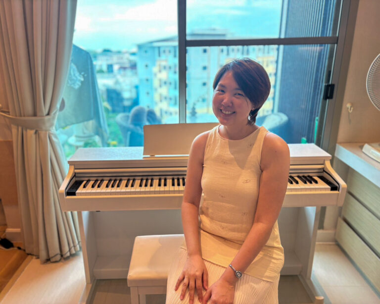 Smiling woman in a cream sleeveless dress sits at a white upright piano in a bright room by a large window.