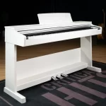 White upright piano on a matching white stand in a studio, with three pedals visible at the base ขายราคาพิเศษ