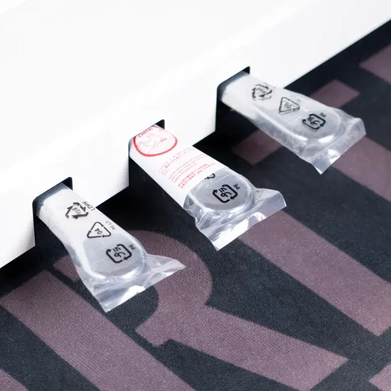 Three individually wrapped disposable items mounted on a white surface, each in a clear plastic pouch above a dark striped carpet. ขายราคาพิเศษ