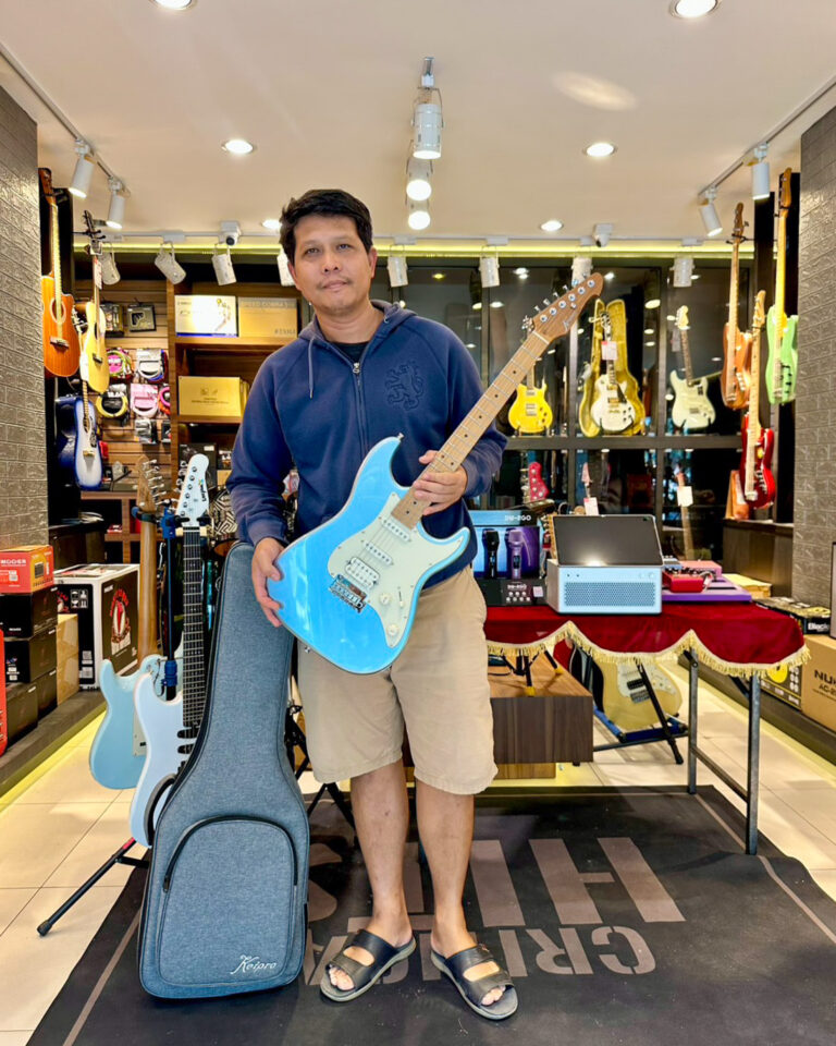 Man in a guitar shop holding a bright blue electric guitar among guitars and gear on display.