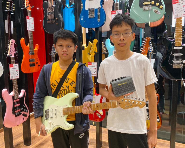 Two boys stand in a guitar shop, one holding a mint-green electric guitar while the other holds a small gray portable amp, with guitars on display behind them.