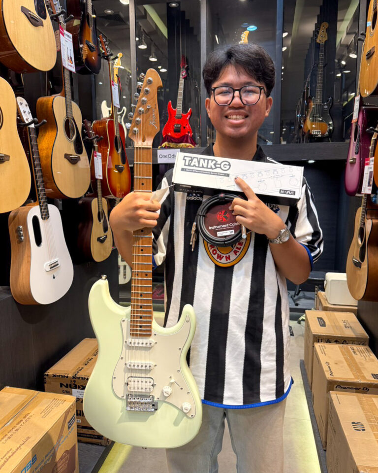 Smiling young man in a guitar shop holds a pale green electric guitar and a boxed accessory, surrounded by guitars on display.