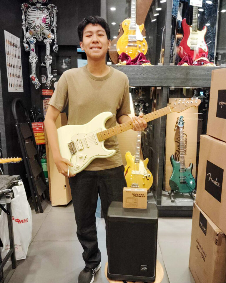 Young man in a beige T-shirt holding a white electric guitar in a guitar shop, with guitars on display behind him and a speaker in front.