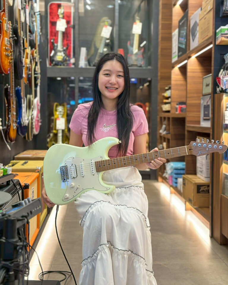 Smiling young woman in a pink t-shirt holding a mint-green electric guitar in a guitar shop.