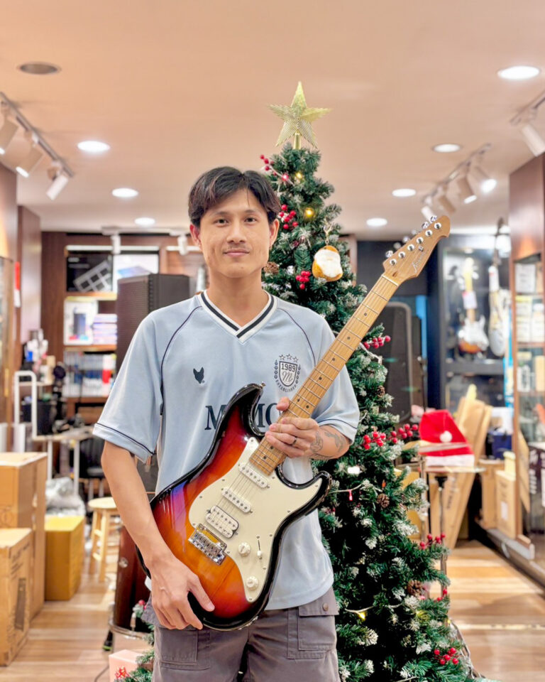 Man wearing a light blue jersey poses with a sunburst electric guitar in a music store beside a Christmas tree behind him.