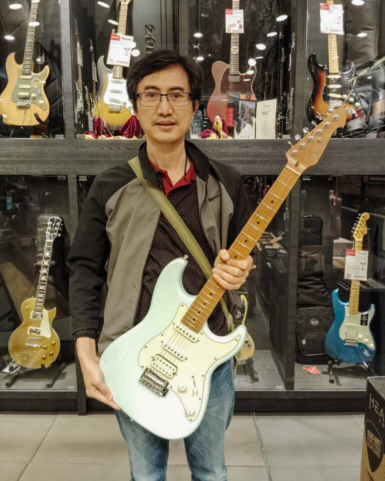 Man in a guitar shop holding a mint-green electric guitar with a white pickguard, standing in front of guitar displays.
