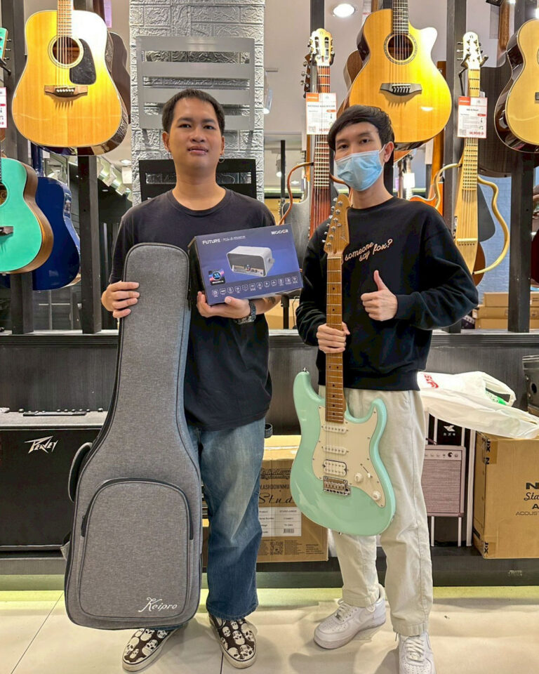 Two men stand in a guitar store; one holds a grey guitar case and a boxed device, the other shows a mint-green electric guitar and wears a face mask, with guitars on the wall behind them.