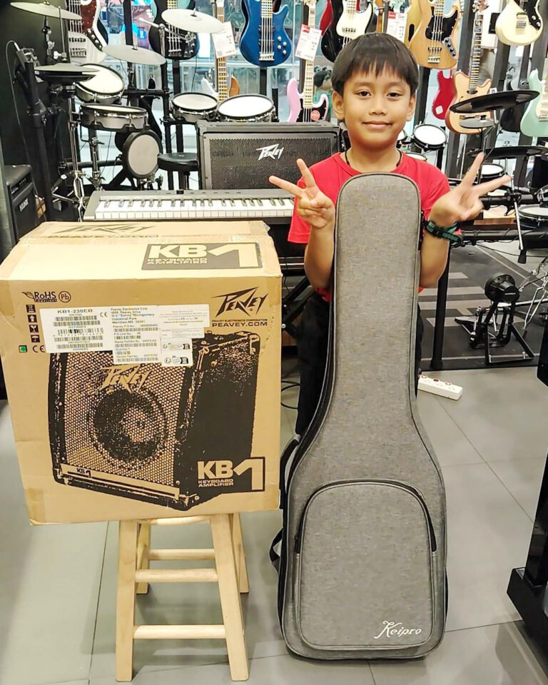 Smiling boy in a red shirt stands beside a boxed Peavey KB1 keyboard amplifier and a gray padded instrument case in a music shop with guitars on the wall behind him.