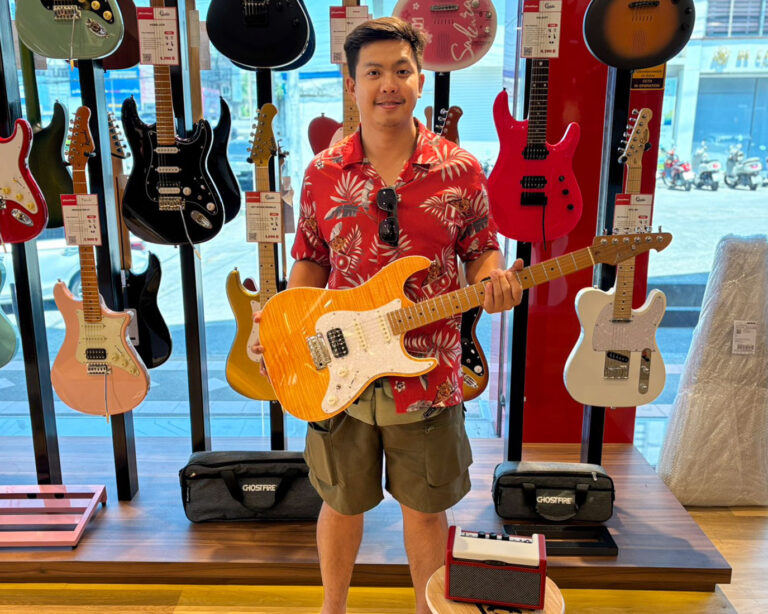 Smiling man in a red Hawaiian shirt holds an orange sunburst electric guitar in a guitar shop with guitars on display behind him.