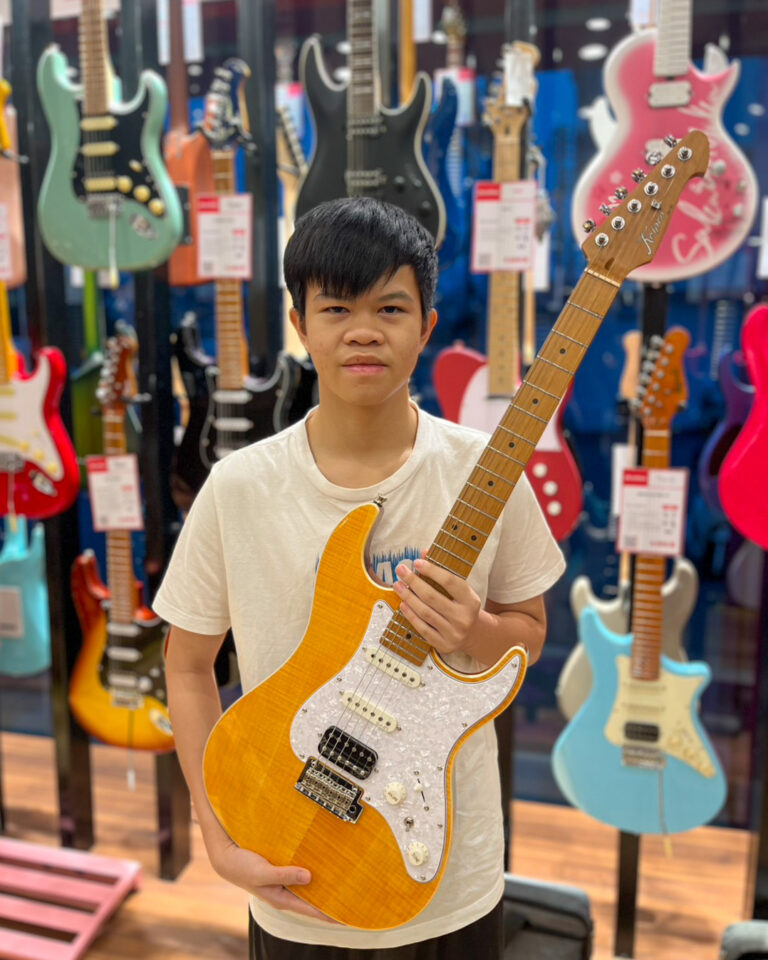 Young boy in a white shirt holds a yellow-orange electric guitar in a guitar shop filled with colorful guitars behind him.