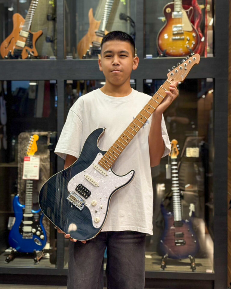 Young man in a white t-shirt posing with a dark blue Fender-style Stratocaster in a guitar shop showroom.