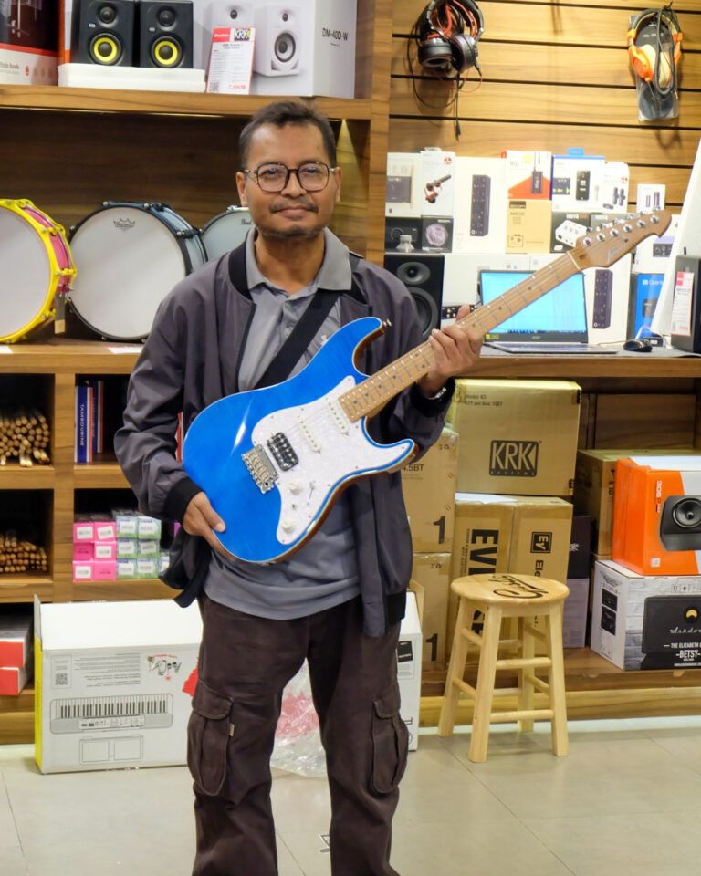 Man in a gray jacket holding a bright blue electric guitar in a music store with speakers and boxes in the background, smiling at the camera.