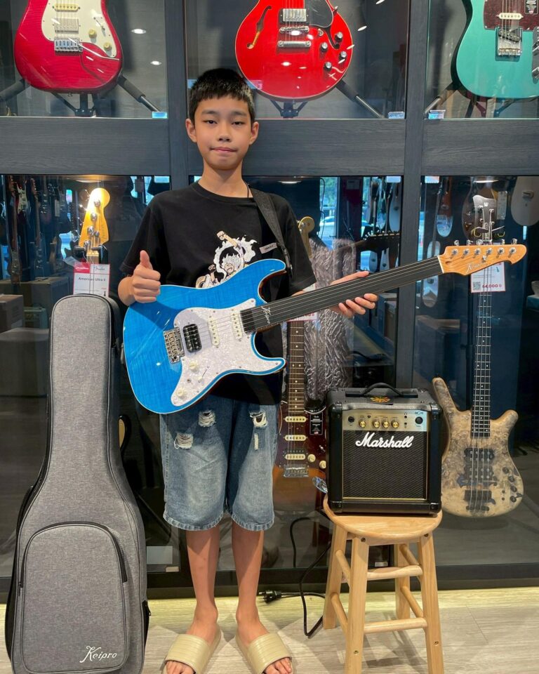 Young boy holding a blue electric guitar in a guitar shop, with amps and guitars on display behind him.