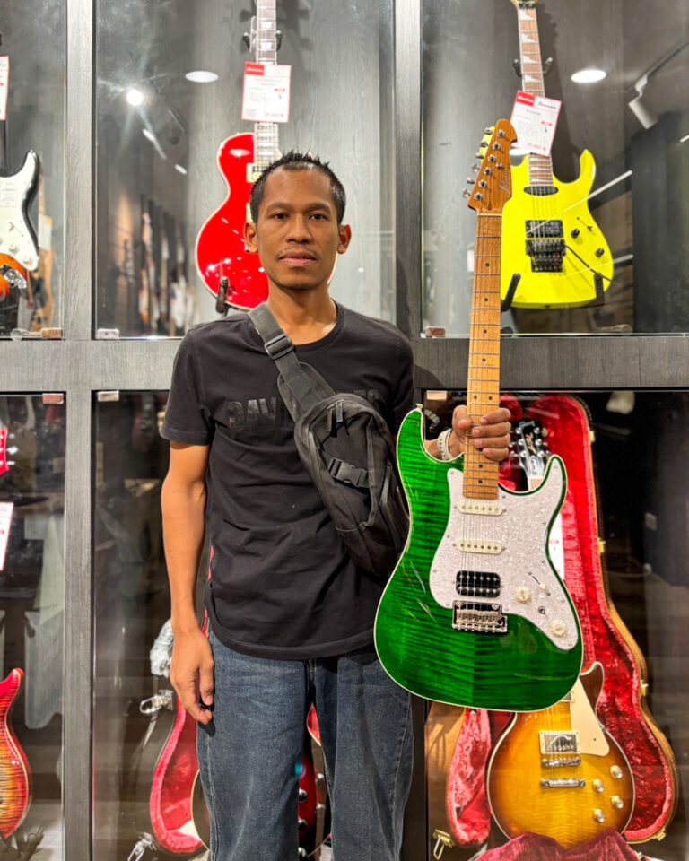Man in a black t-shirt and jeans holding a vibrant green electric guitar with a white pickguard in a guitar shop display case.