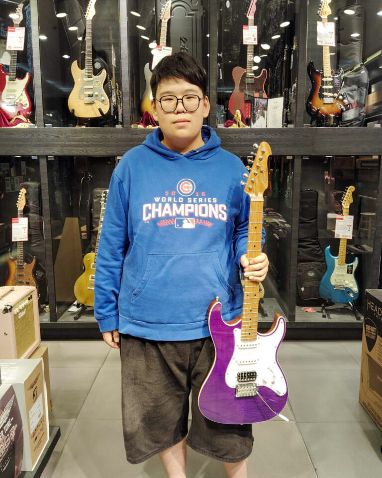 Young boy with glasses in a blue hoodie stands in a guitar shop, holding a purple electric guitar.