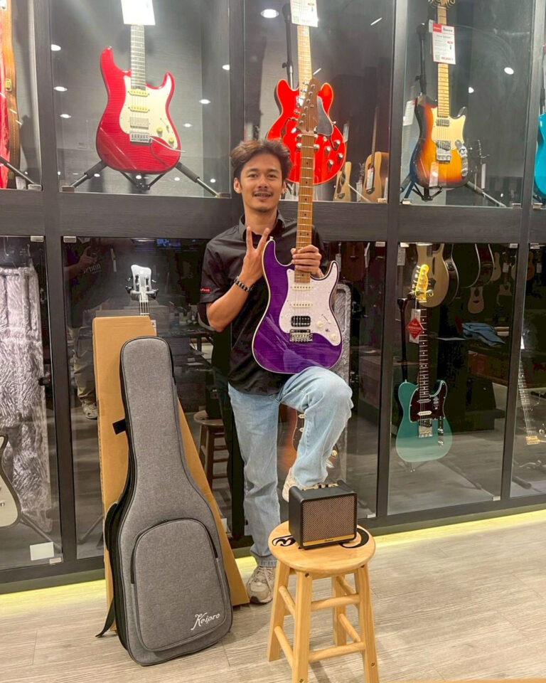 Person in a guitar shop holding a purple electric guitar with white pickguard, standing beside guitar cases and a small amp on a wooden stool.