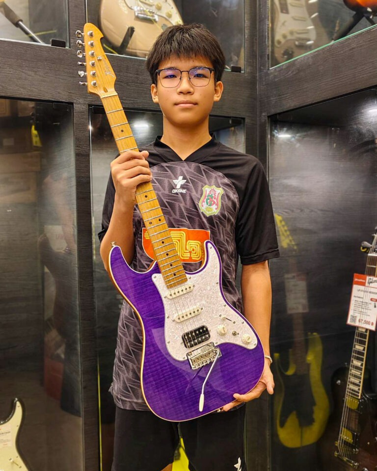 Young boy with glasses holding a purple electric guitar in a guitar shop, facing the camera.