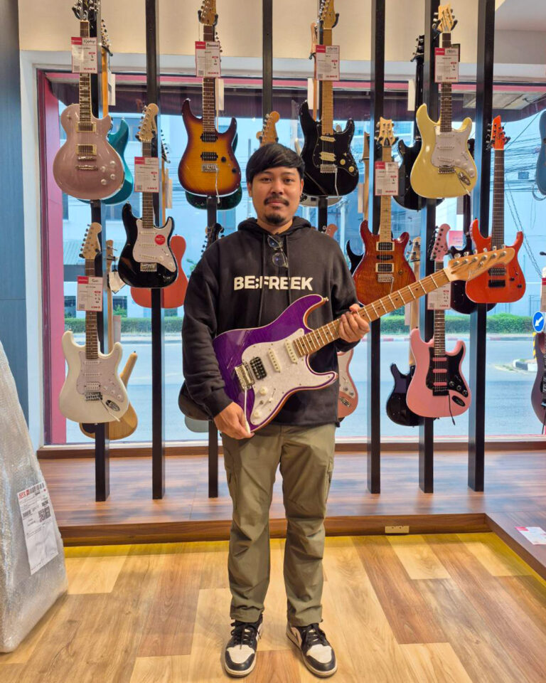 Man stands in a guitar shop holding a purple electric guitar with a white pickguard, shelves of guitars behind him.