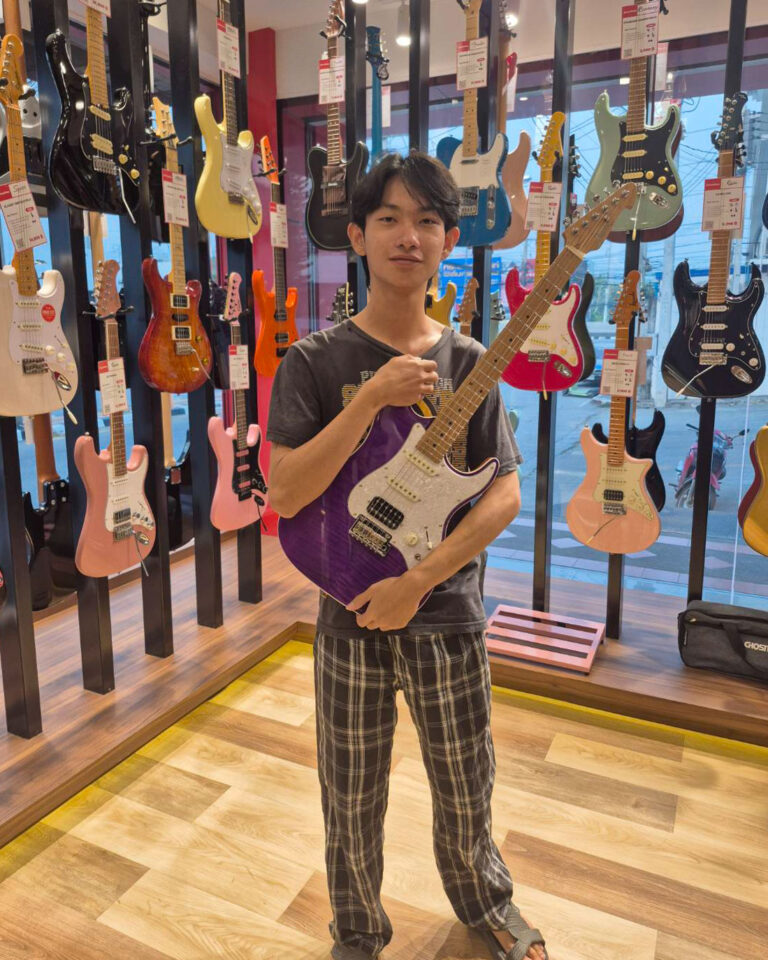 Young man standing in a guitar shop holding a purple electric guitar.
