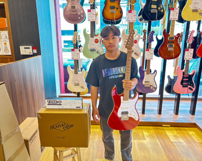 Young man in a guitar shop holding a red electric guitar in front of a wall of colorful guitars on display.