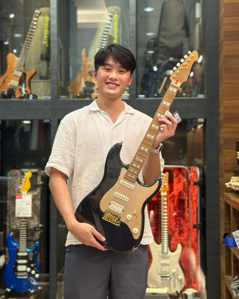 Smiling young man in a light shirt poses with a black electric guitar inside a guitar shop, shelves of guitars visible in the background.