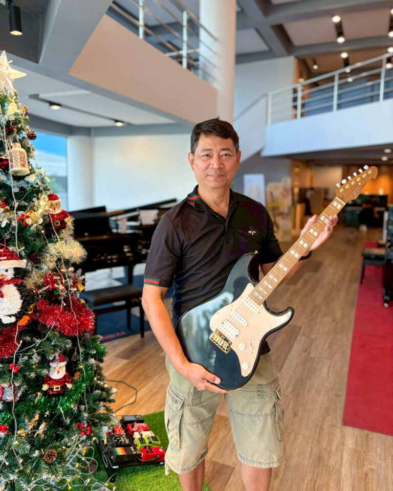 Man in black polo and khaki shorts holding an electric guitar in a showroom next to a decorated Christmas tree.