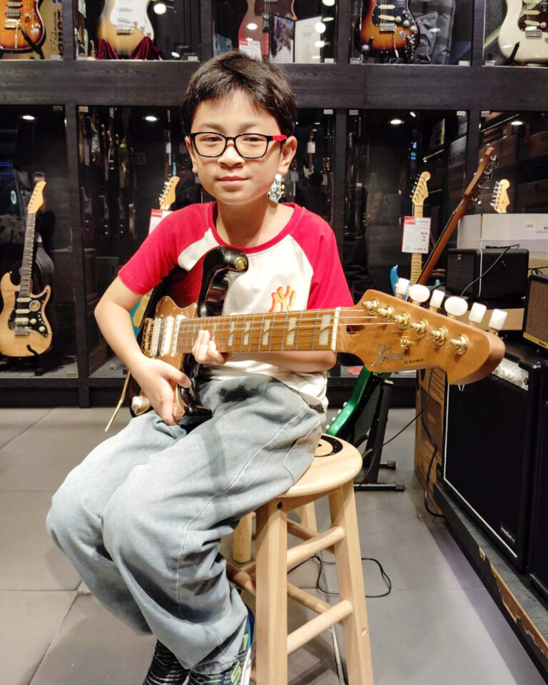 A young boy in glasses sits on a wooden stool, playing an electric guitar in a guitar shop with guitars on display behind him.