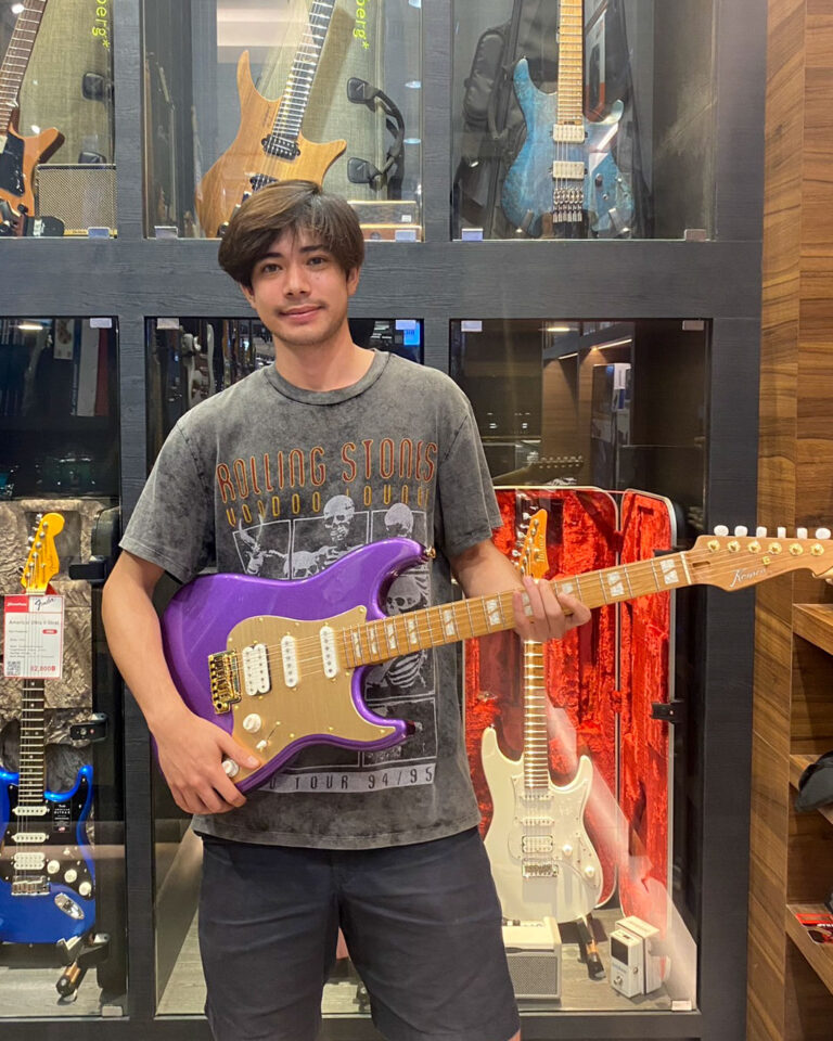 Man in a gray Rolling Stones t-shirt posing with a purple electric guitar in a guitar shop display wall.