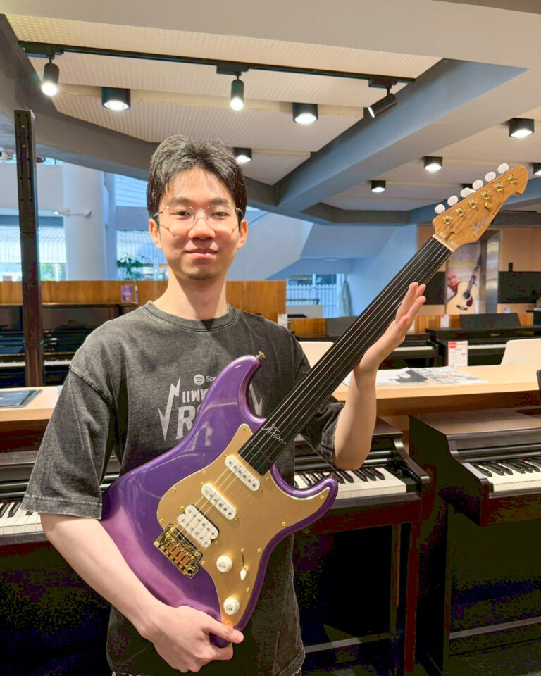 Young man with glasses holds a purple Stratocaster-style electric guitar in a piano showroom with pianos in the background.