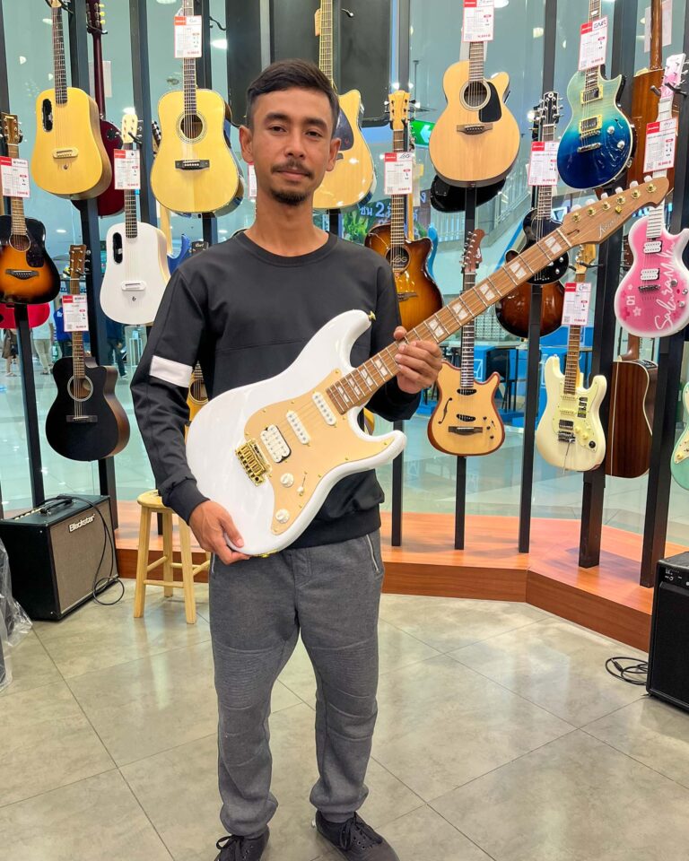 Man in a dark long-sleeve shirt holding a white electric guitar in a guitar shop with many guitars on display behind him.