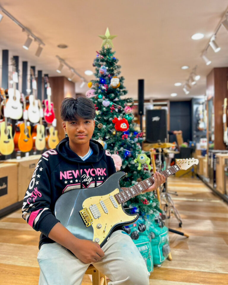 Young man sits with a black electric guitar in a guitar shop decorated for Christmas near a Christmas tree.