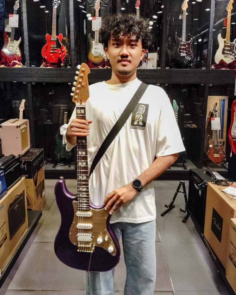 Person in a white T-shirt holding a purple electric guitar with a strap in a guitar shop.