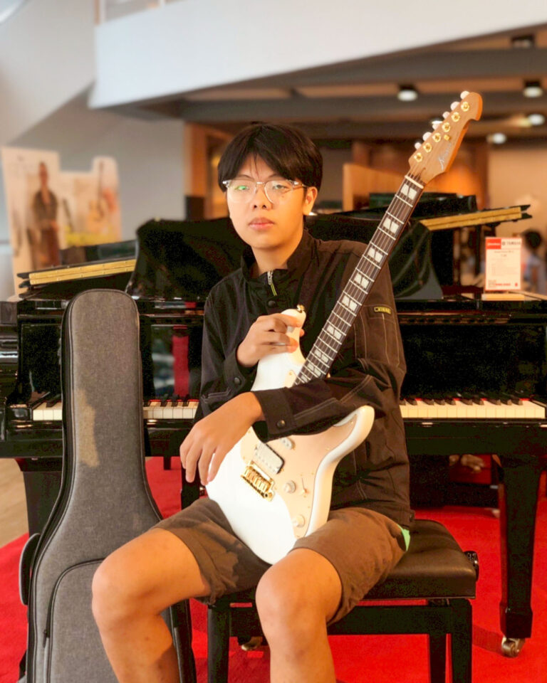 Young person with glasses holding a white electric guitar in a music room, seated in front of a piano.