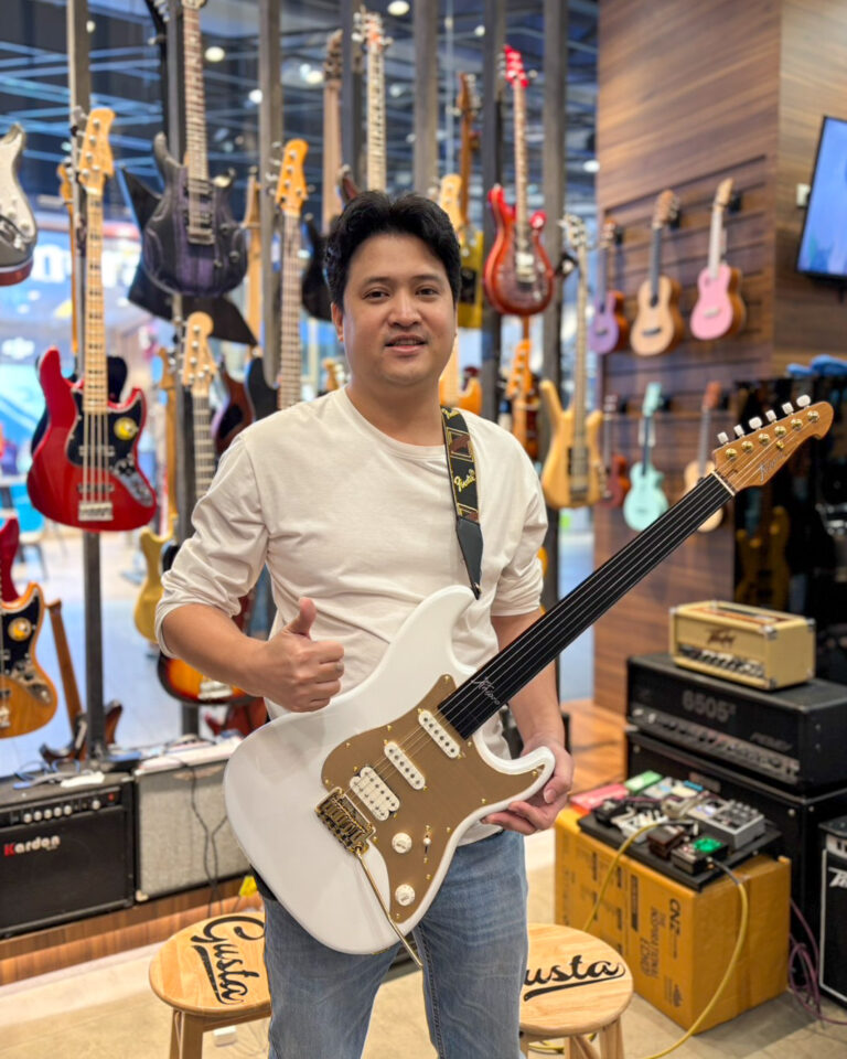 Smiling man in a white shirt holding a white electric guitar in a guitar shop, surrounded by hanging guitars