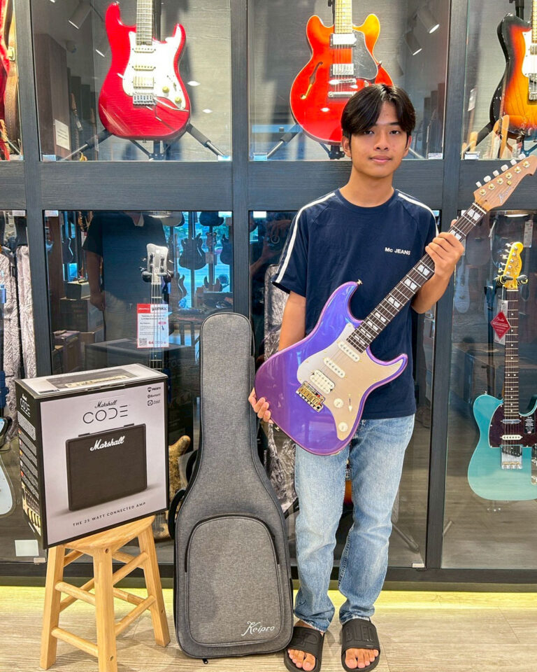 Young man in a guitar shop holding a purple electric guitar in front of display cases filled with guitars and accessories.
