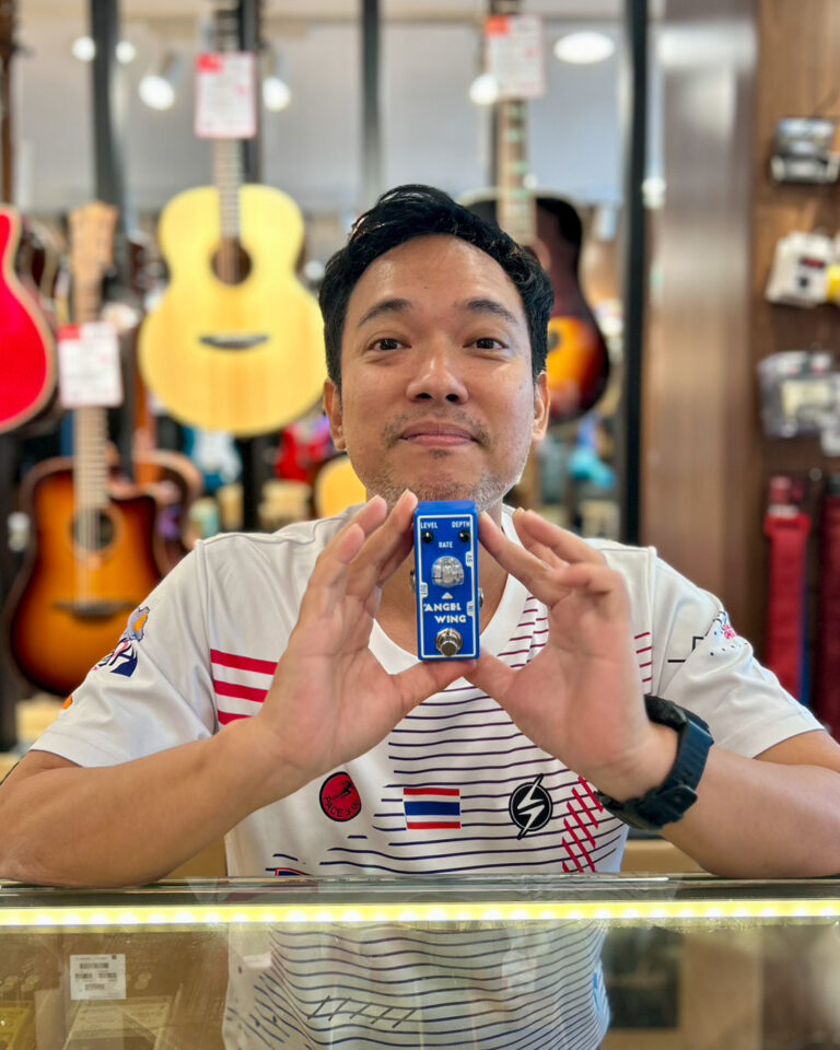 Man in a guitar shop holds up a blue Angel Wing guitar effects pedal toward the camera.