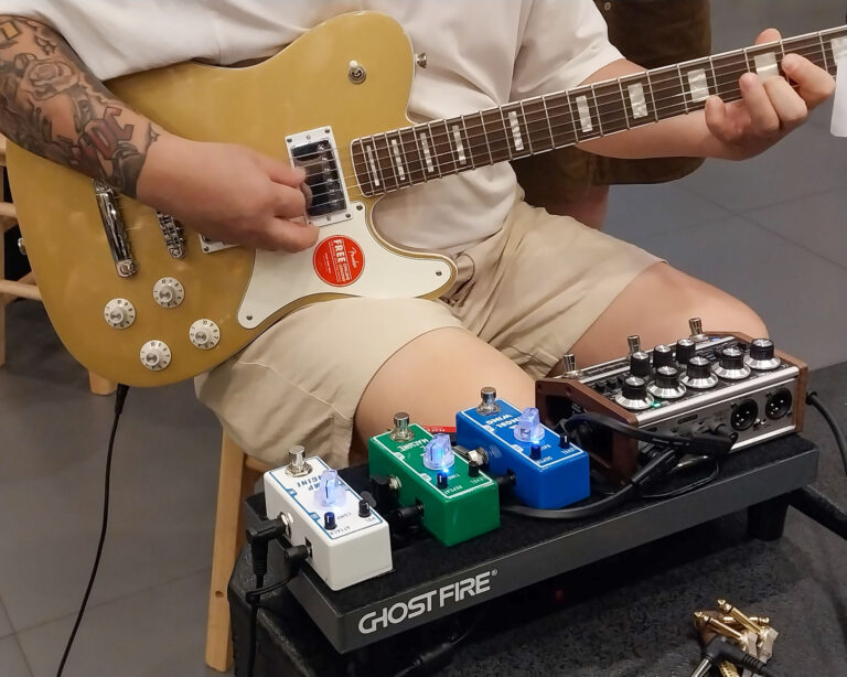 Person playing a gold electric guitar with a cream pickguard, tattooed left forearm visible, on a pedalboard setup nearby.